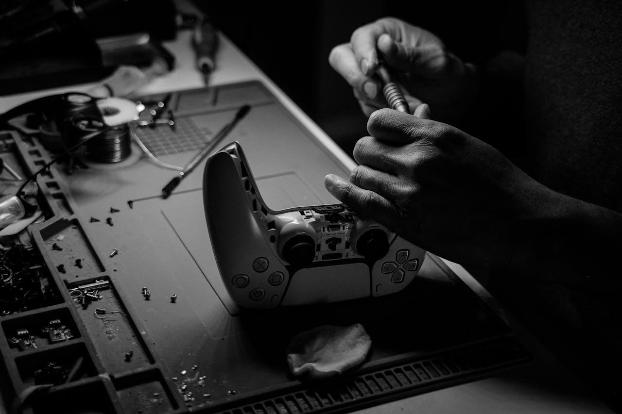Close-up black and white image showing hands repairing a game controller at a workbench.